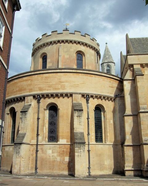 Architecture. A round church made of cream coloured stone is tucked between other buildings. Engaged buttresses alternate with narrow, arched windows. The narrower, also circular, second storey has crenelations around the top edge.