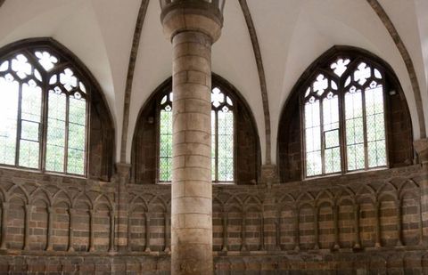 Architecture. A pillar rises from the centre of this meeting room. The walls we can see are lined with stone seats under blind, overlapping arches, with three large windows above.