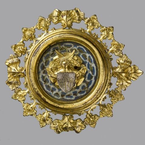 Plaque. A golden, medallion-like roundel has a gold angel with crossed wings and a silver-coloured shield at its centre, against a background enamelled in blue with a white figure-of-eight pattern. The perimeter is lined with flaring gold leaves.