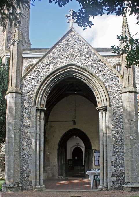 Architecture. A covered porch projects from the side of a grey stone church. The opening is a pointed arch set under a triangular, peaked stone wall flanked by spires.