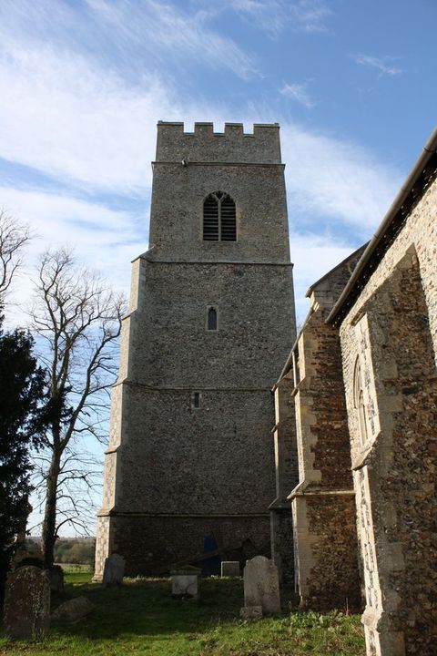Architecture. A four-storey, wide stone tower rises over a porch projecting from the side of a church. In this view, the right edge, facing the church, is straight, and the front left corner has stepped buttresses. Small windows pierce the second and third levels, and a larger opening is on the top storey, which is crenelated.